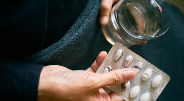 Dłoń trzyma blister z tabletkami i szklankę wody. Czy to próba zwalczenia nadmiaru witaminy B12?